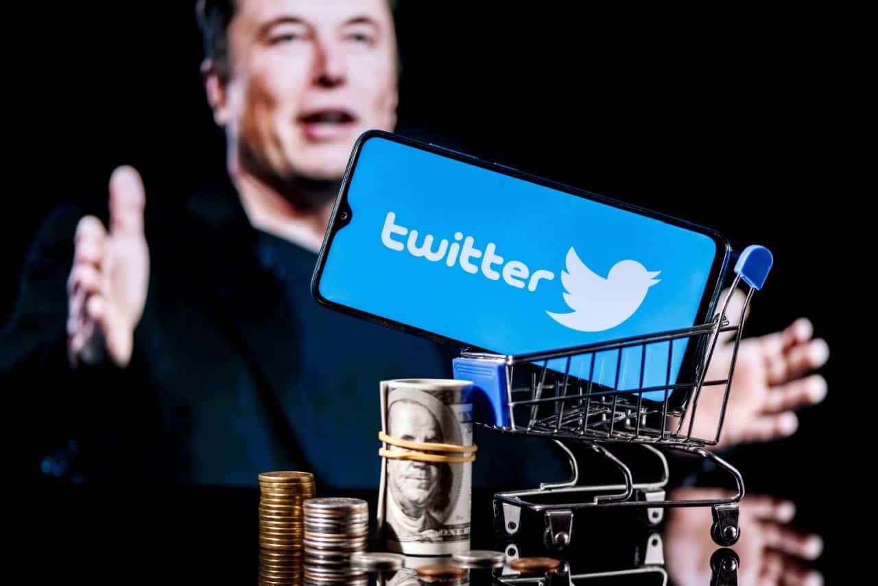 Celular mostrando logo do Twitter dentro de um carrinho de compras em miniatura. Ao lado, várias moedas e um rolo de notas de dólar. Ao fundo, imagem de Elon Musk de braços abertos desfocada.