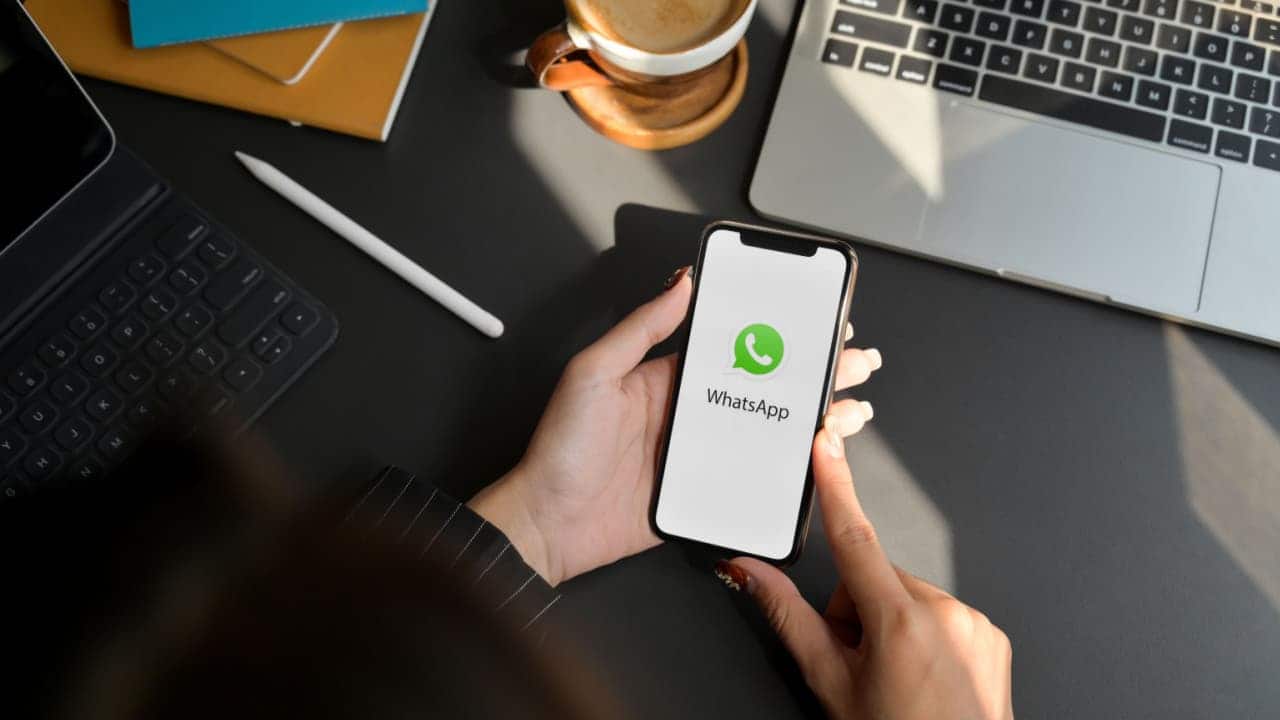 Mãos de uma pessoa segurando celular que mostra logo do WhatsApp. As mãos estão apoiadas em uma mesa de escritório, com computador, caneta e outros itens comuns de empresas.