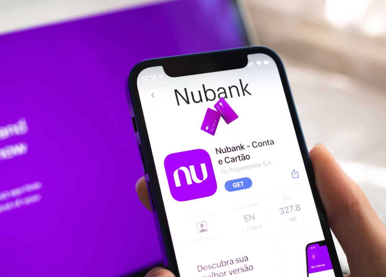 Mão segurando celular com a tela inicial do aplicativo do Nubank. Fundo roxo desfocado