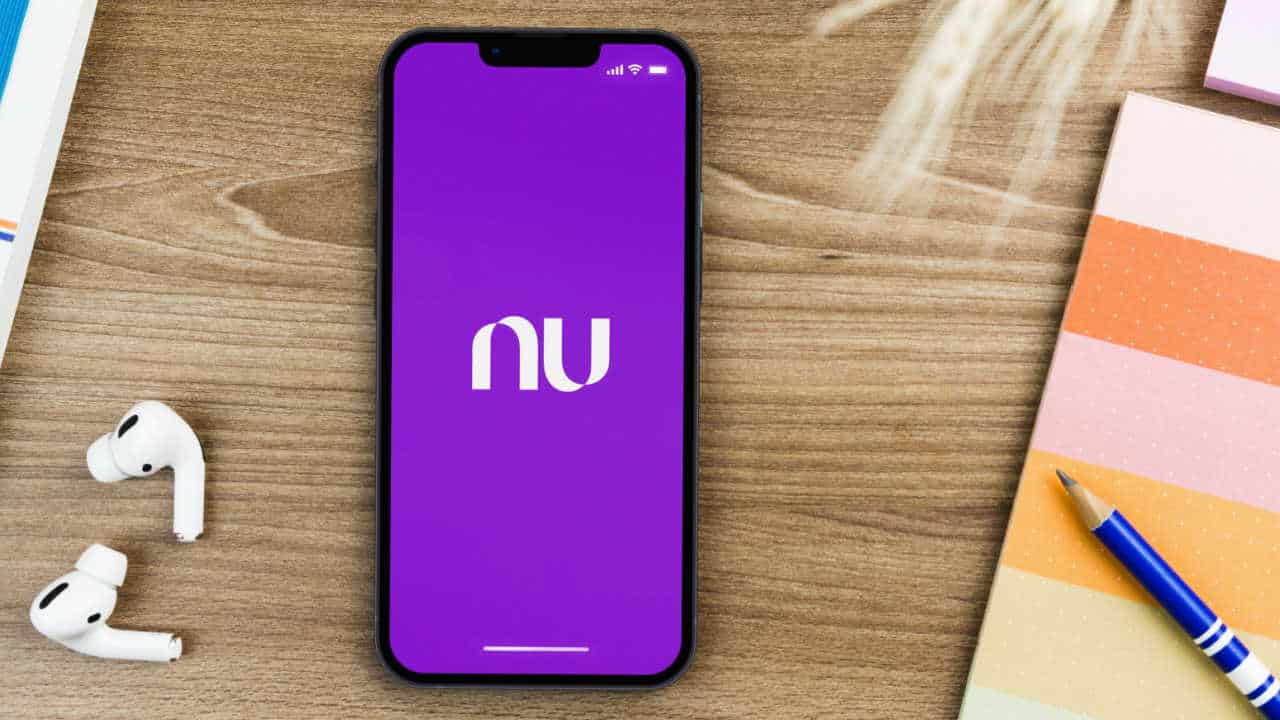 Imagem de celular com app do Nubank na tela inicial. Ao lado do aparelho tem fones de ouvido, um caderno e um lápis.
