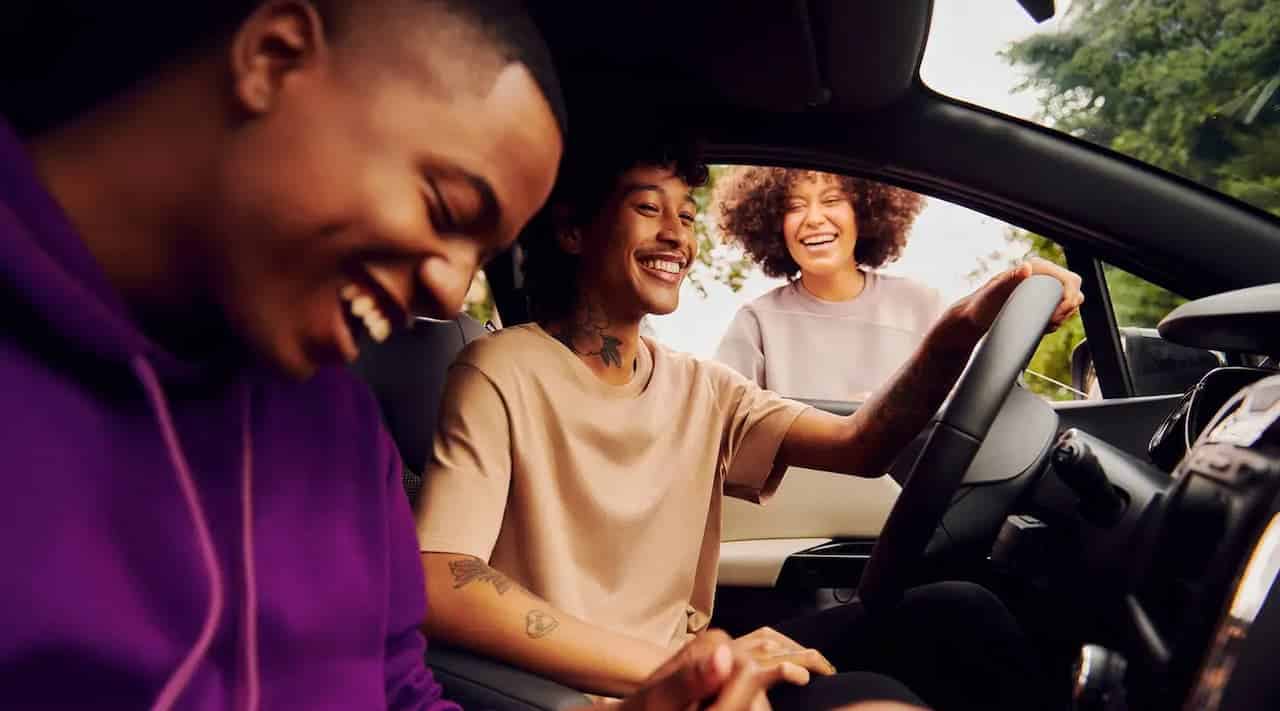 Dois jovens sorrindo dentro do carro enquanto conversam com uma jovem que está na janela do carro