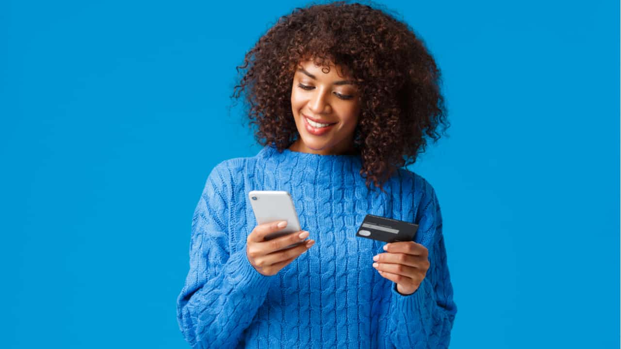 Mulher preta de roupa azul, em fundo azul, com um celular branco em uma mão e um cartão preto em outra. Ela sorri olhando para o celular