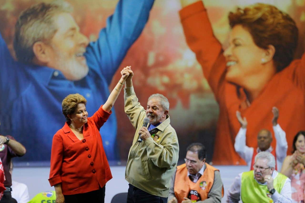 Dilma Rousseff e Luiz Inácio Lula da Silva, em evento político, de mãos dadas e com os braços levantados. Ao fundo, uma grande imagem dos dois fazendo a mesma pose.