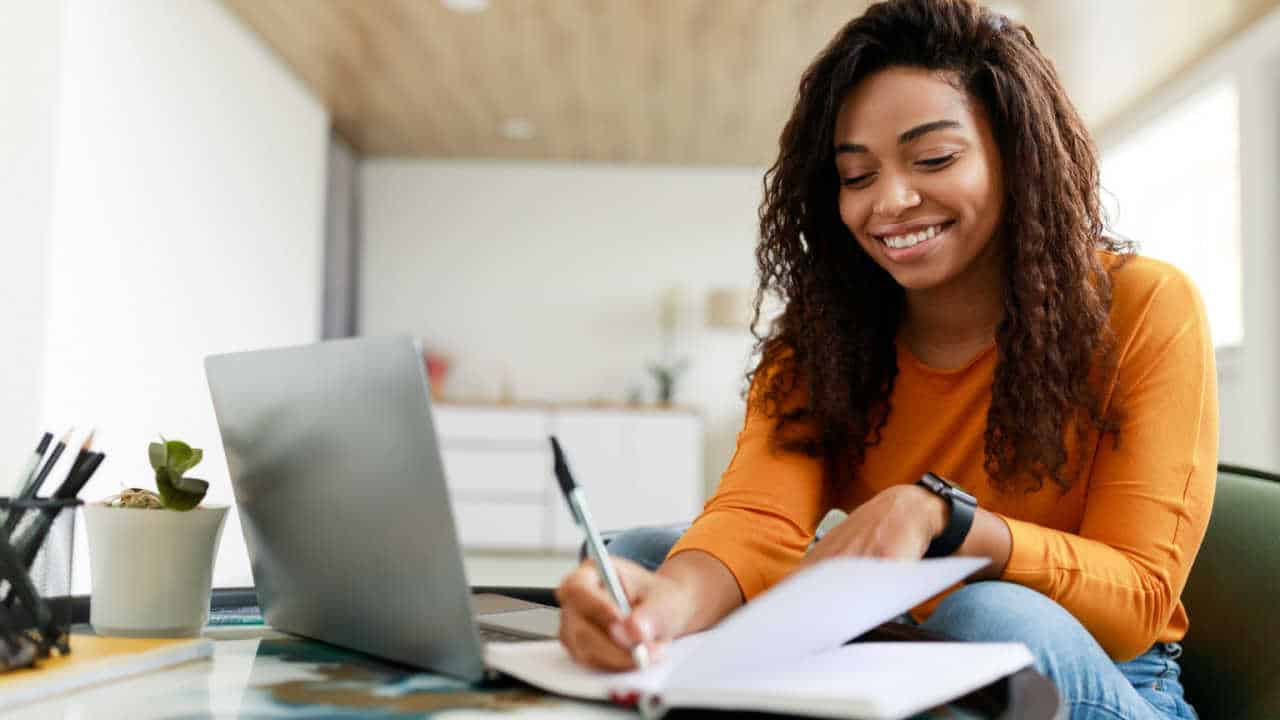 Mulher sorrindo, assinando papéis e com o notebook aberto na mesa, dando a entender que está abrindo seu MEI.