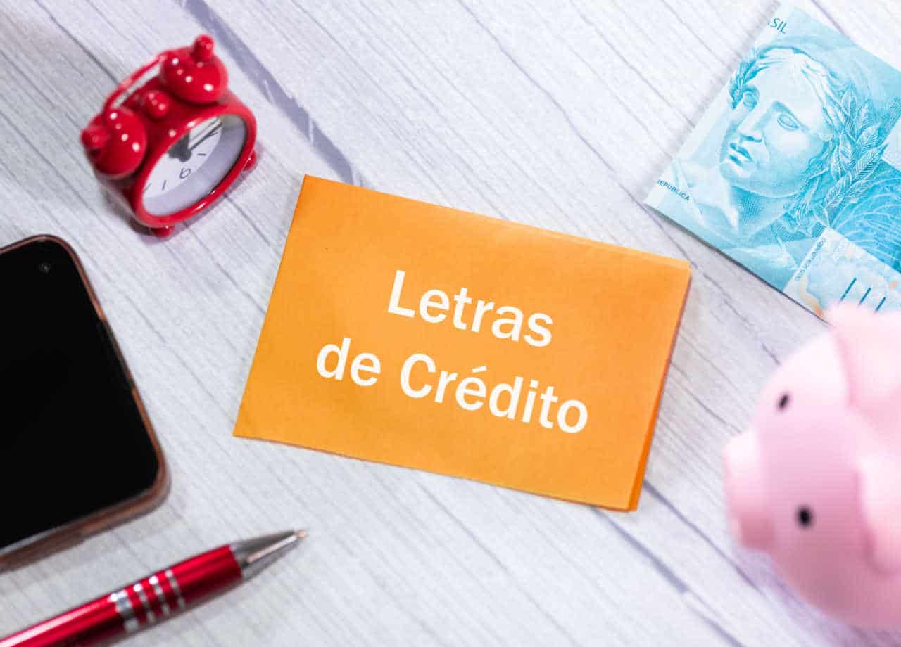 falha de papel laranja escrito "Letras de Crédito" sobre uma superfície plana semelhante a uma mesa. Também nessa superfície estão um despertador analógico, uma caneta vermelha, uma nota de cem reais, um cofre em formato de porquinho e uma carteira preta. LCI