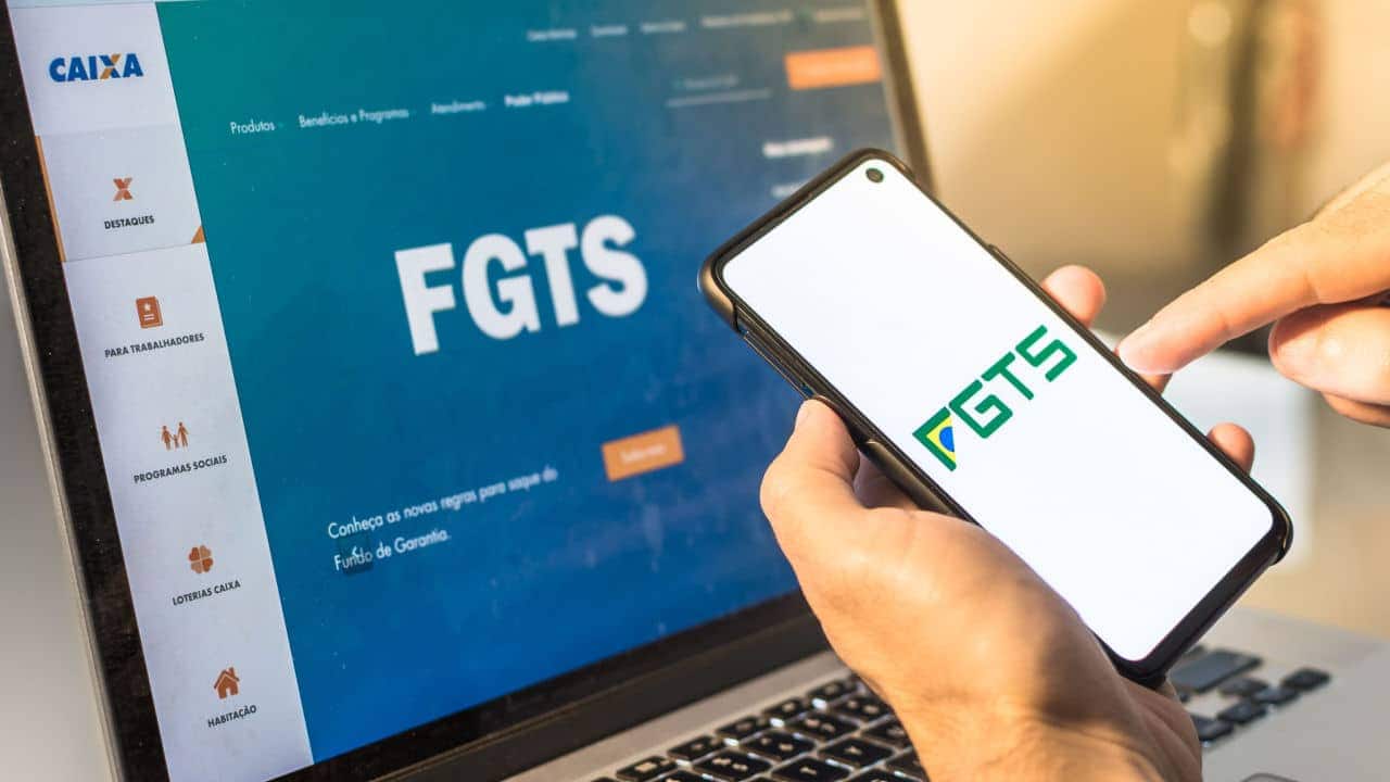 Mão segurando celular com a logo do FGTS, notebook de fundo com a tela inicial no site do FGTS