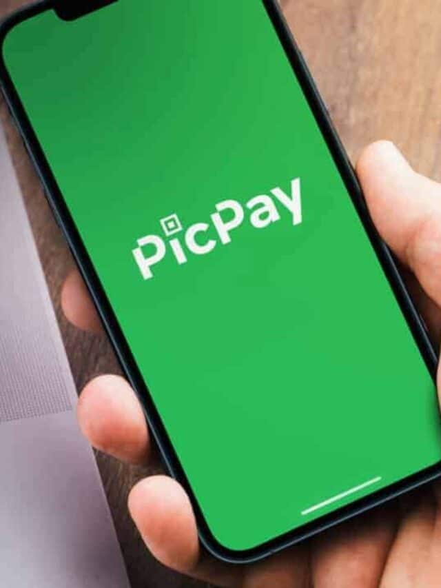 Posicionada ao lado de um notebook, mão segura um celular que está iniciando o app do PicPay