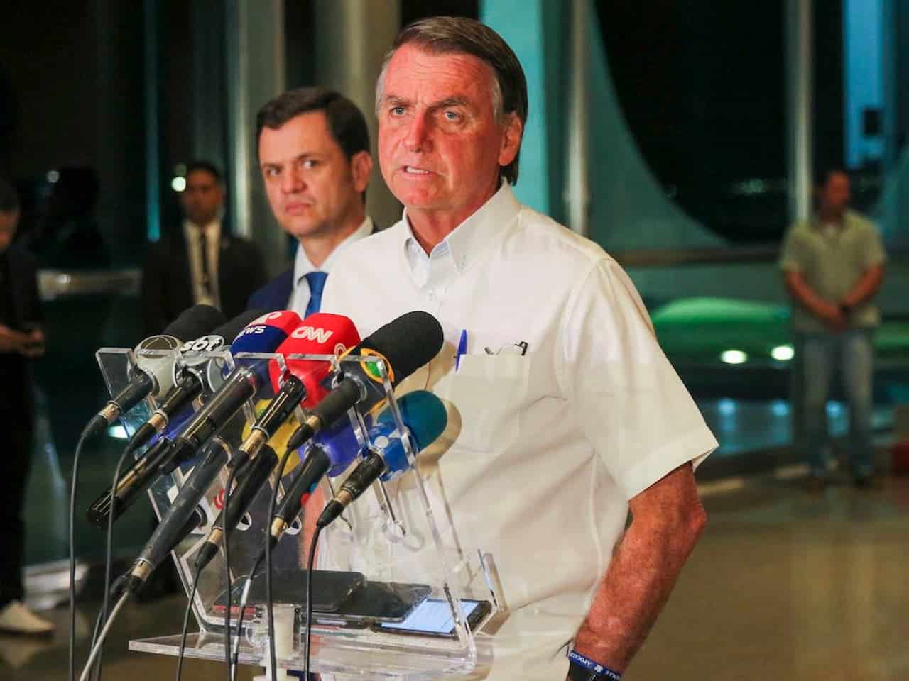 ex-presidente Bolsonaro vestindo camisa branca em frente a diversos microfones de televisão e discursando para a impressa