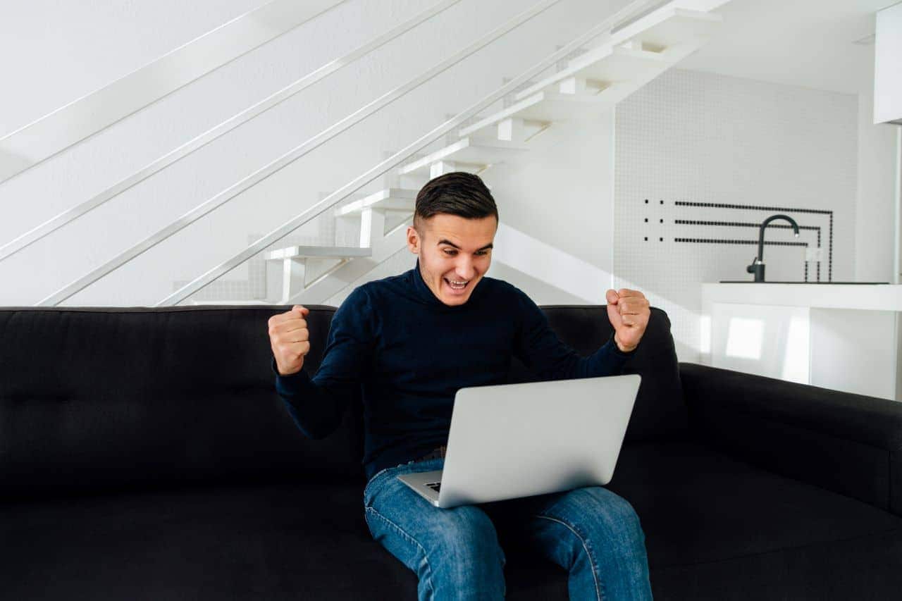 Homem sentado no sofá com notebook no colo e com as mãos para cima em comemoração a um lance de leilão concurso