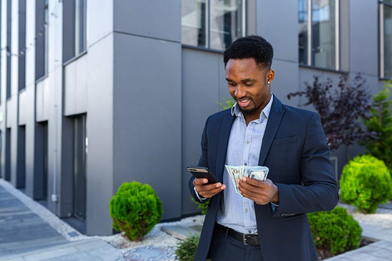 Imagem mostra um homem sorrindo enquanto segura o celular com a mão direita, enquanto segura notas de dinheiro na mão esquerda.