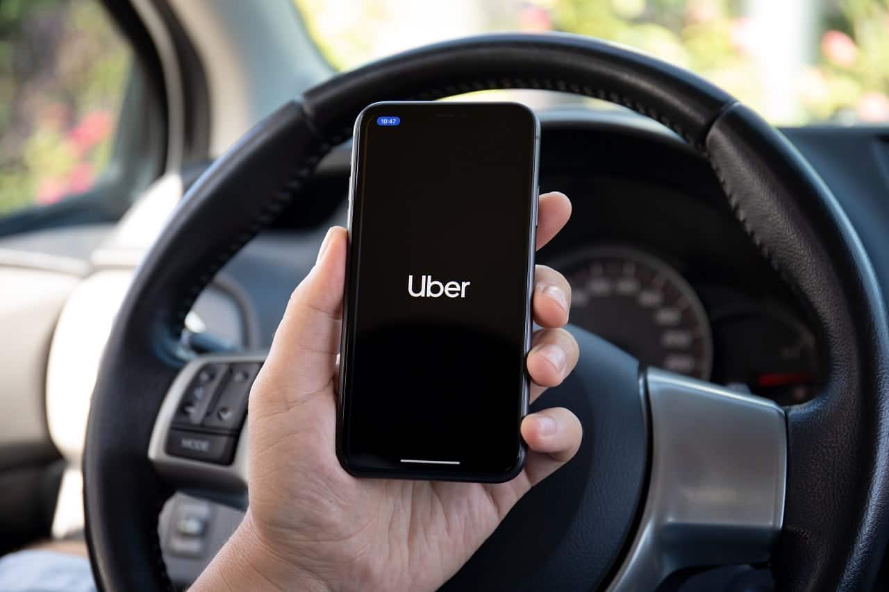 Imagem mostra uma mão masculina segurando um celular com o aplicativo Uber aberto, na frente de um volante.