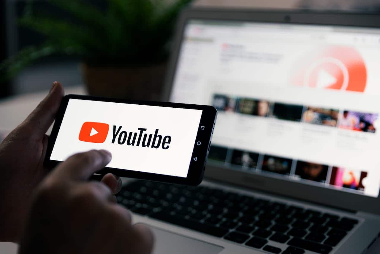 mão segurando smartphone com youtube , ao fundo um laptop com youtube aberto