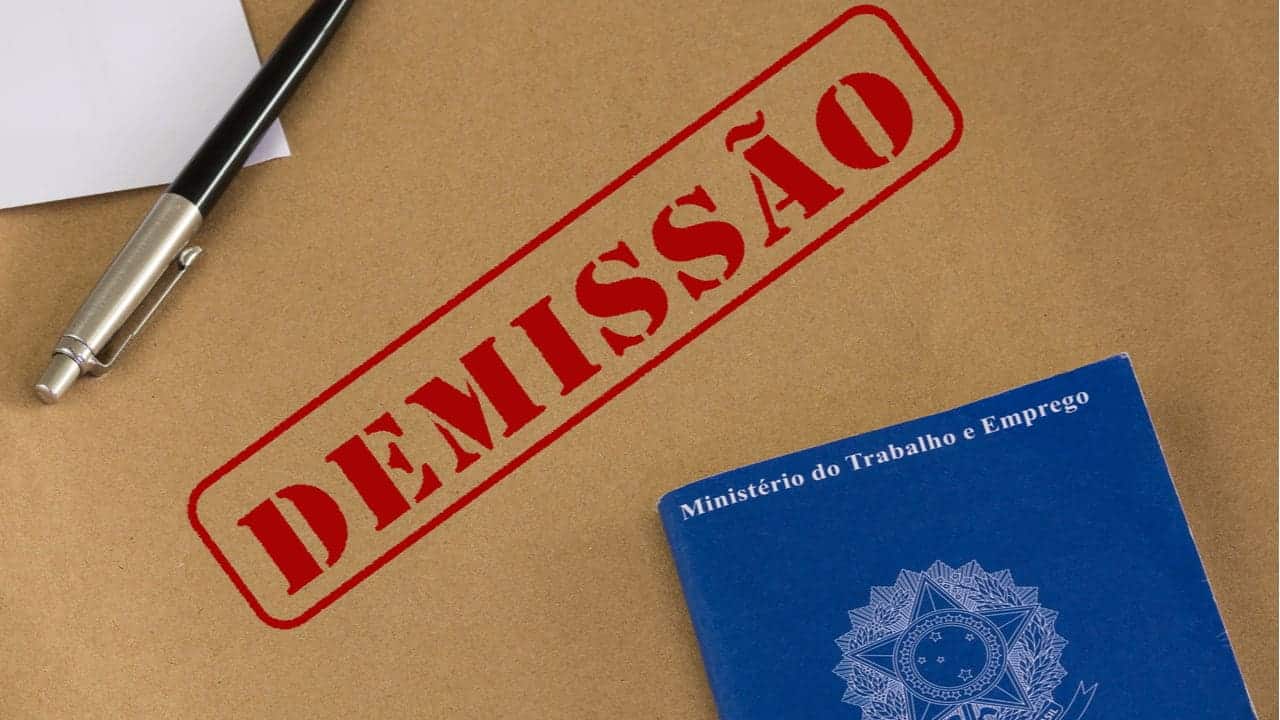 Papelão escrito "demissão" com tinta vermelha, e em cima tem uma carteira de trabalho e uma caneta