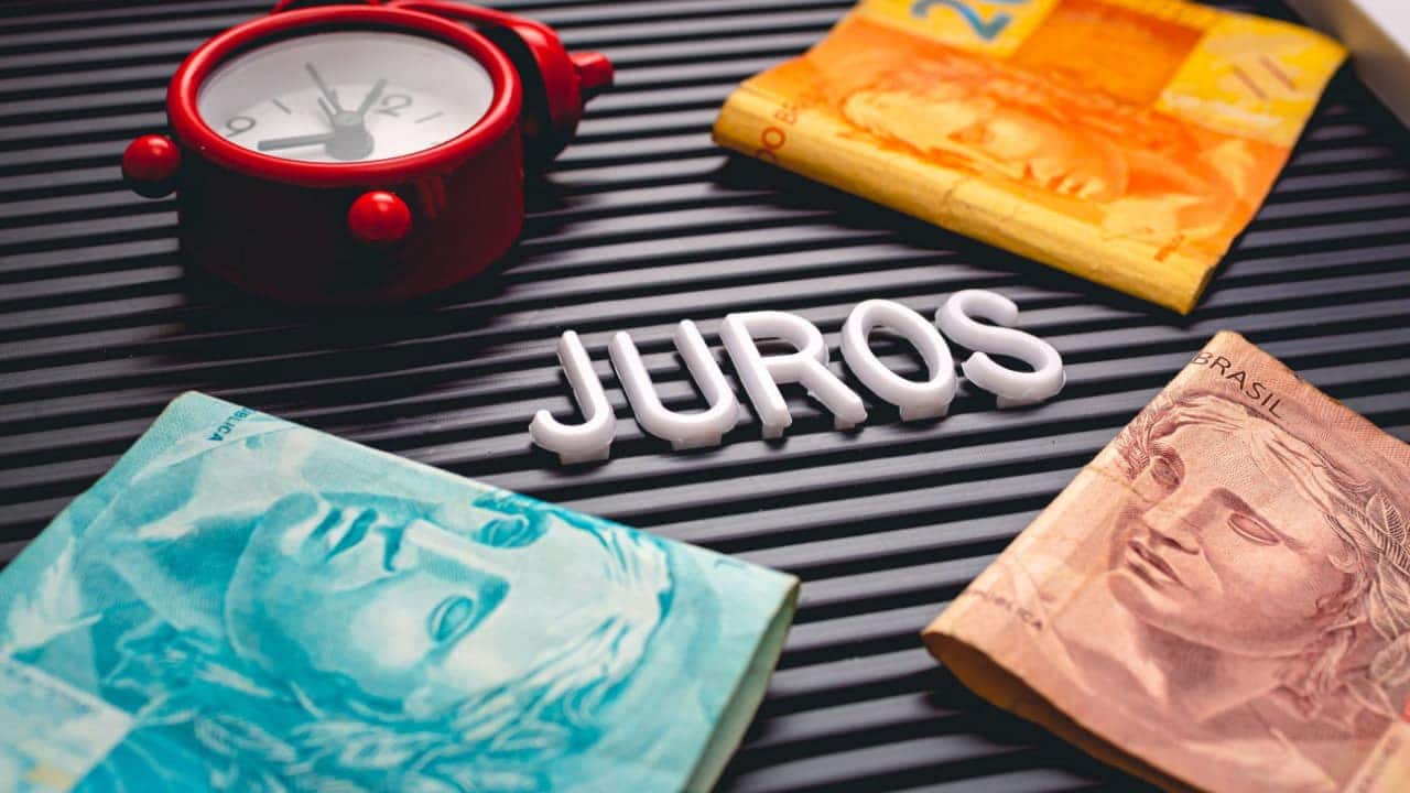 Notas de 10, 20 e 100 reais ao lado de um relógio. Os itens estão sobre uma placa com as letras da palavra juros.