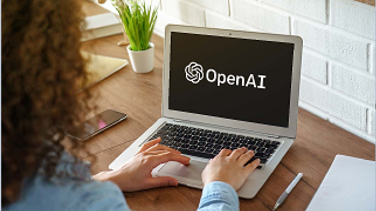 Mulher de cabelos cacheados digitando em seu notebook. Na tela dele, a escrita "OpenAI" sobreposta em um fundo preto.