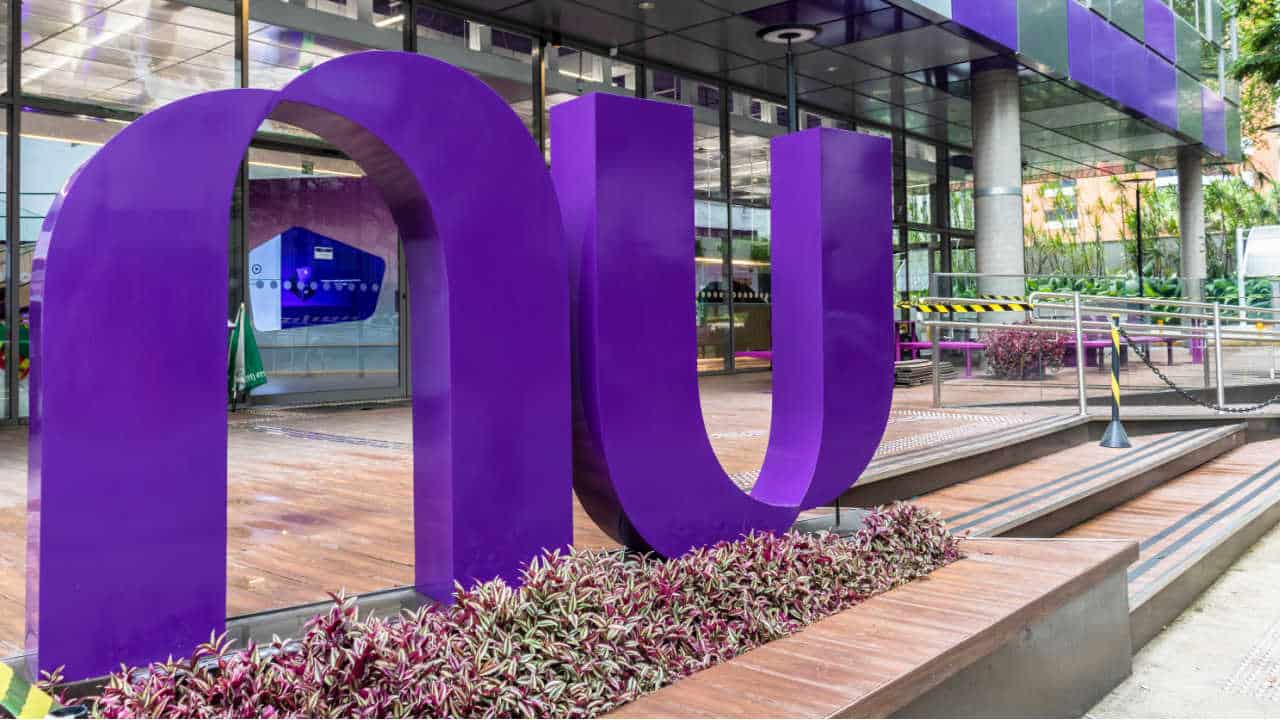 Fachada de uma unidade do Nubank com letreiro roxo disposto horizontalmente em frente a um prédio