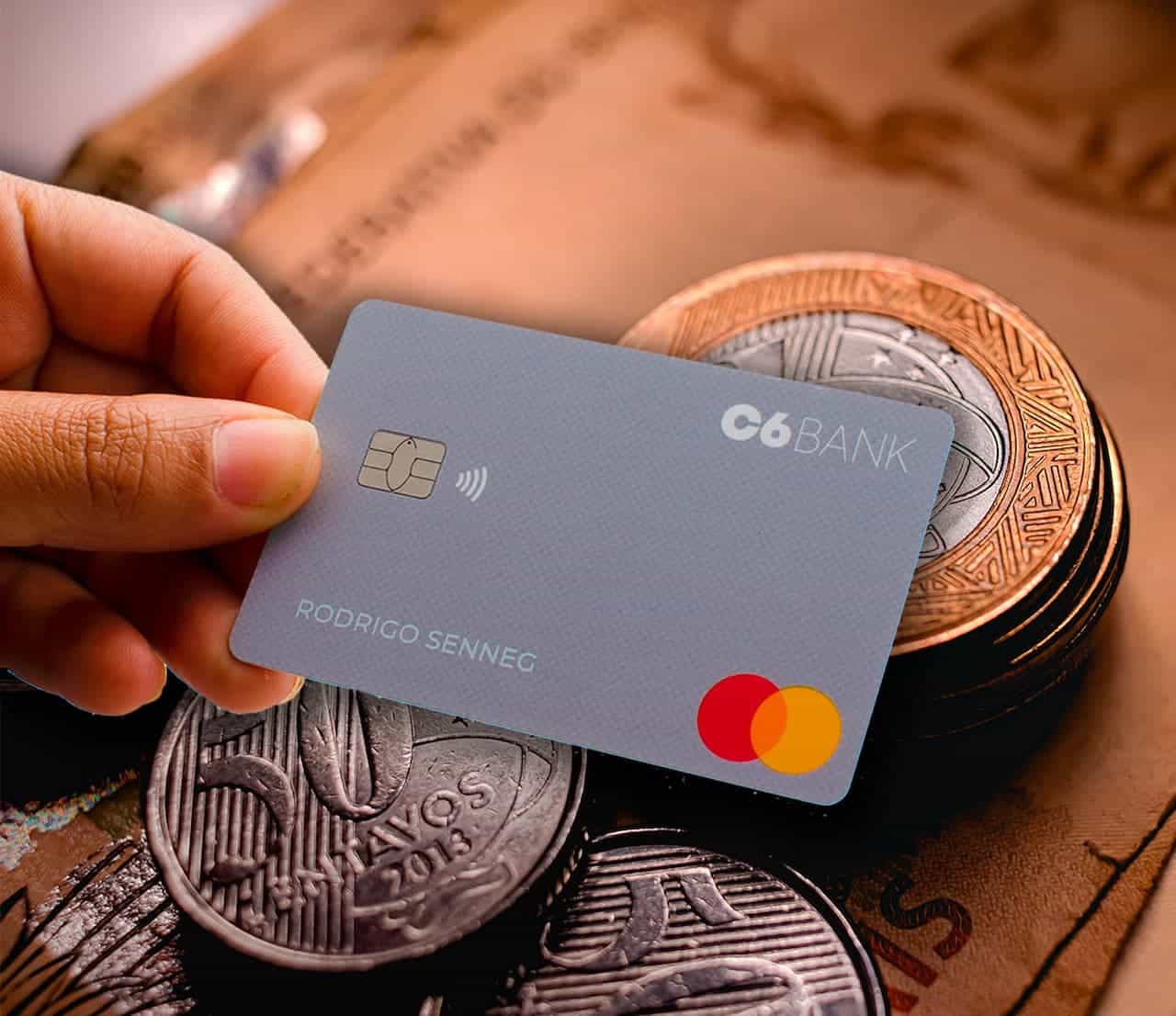 Pessoa segurando cartão do C6 Bank com notas de dinheiro e moedas ao fundo
