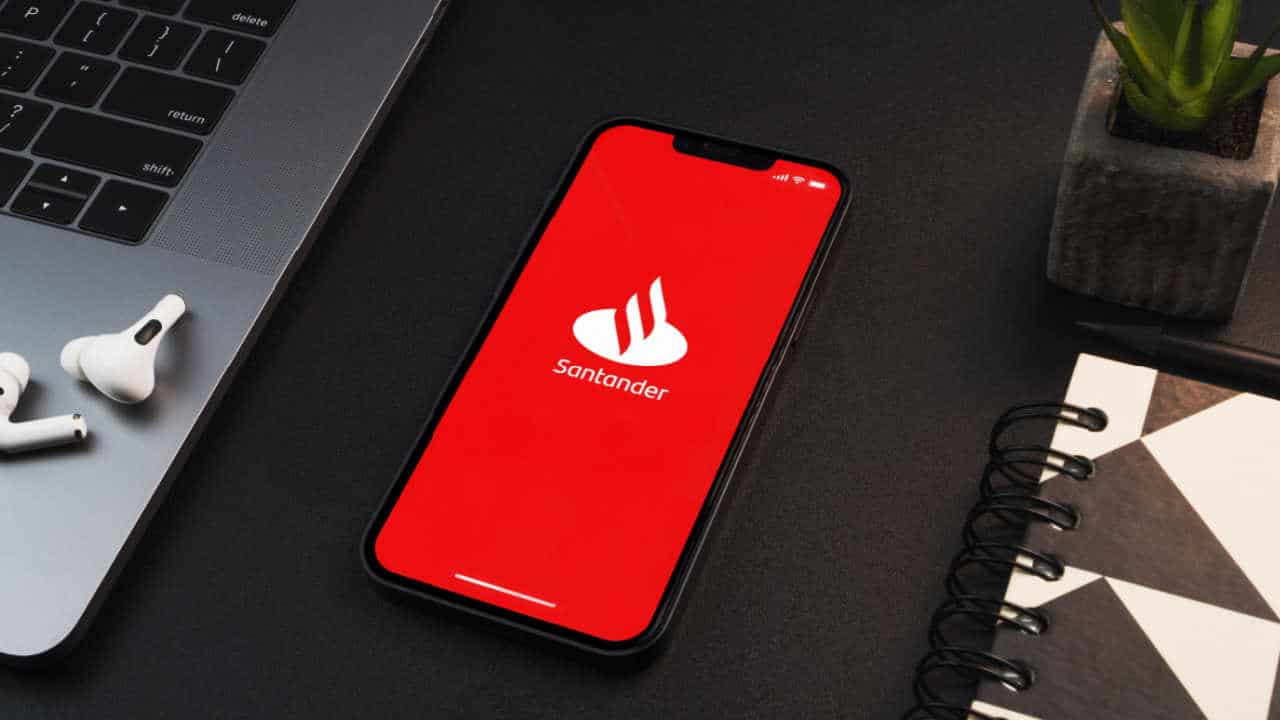 celular com app do Santander na tela inicial. O aparelho está sobre uma mesa, ao lado de um notebook, fones de ouvido e um caderno.