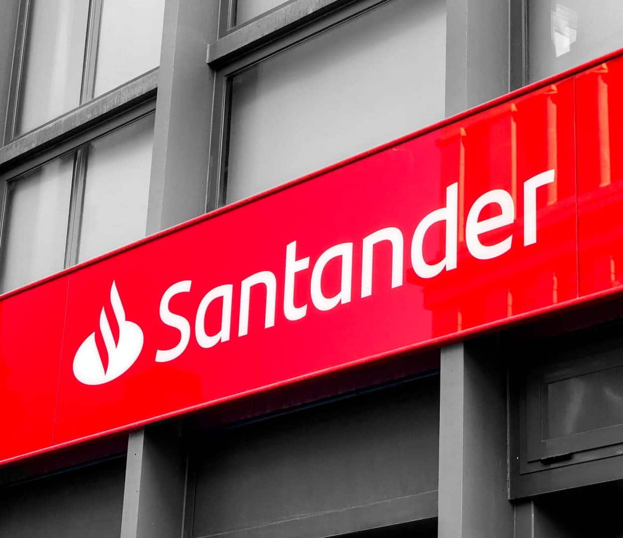 Fachada com placa do Santander em branco e vermelho. Como suporte da fachada, prédio cinza ao fundo