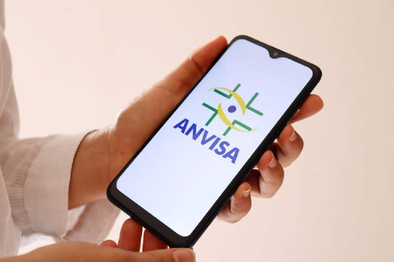 Uma mão segurando um celular com o logo da Anvisa na tela