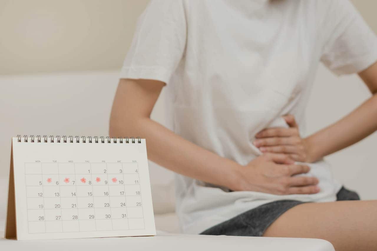 Calendário com dias marcados. Ao fundo uma pessoa com cólica. Imagem faz referência à licença menstrual