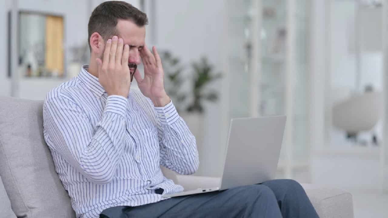 Um homem sentado em um sofá com o notebook sobre as pernas. Ele leva as mãos a cabeça como se estivesse sofrendo, enquanto olha para a tela do computador.