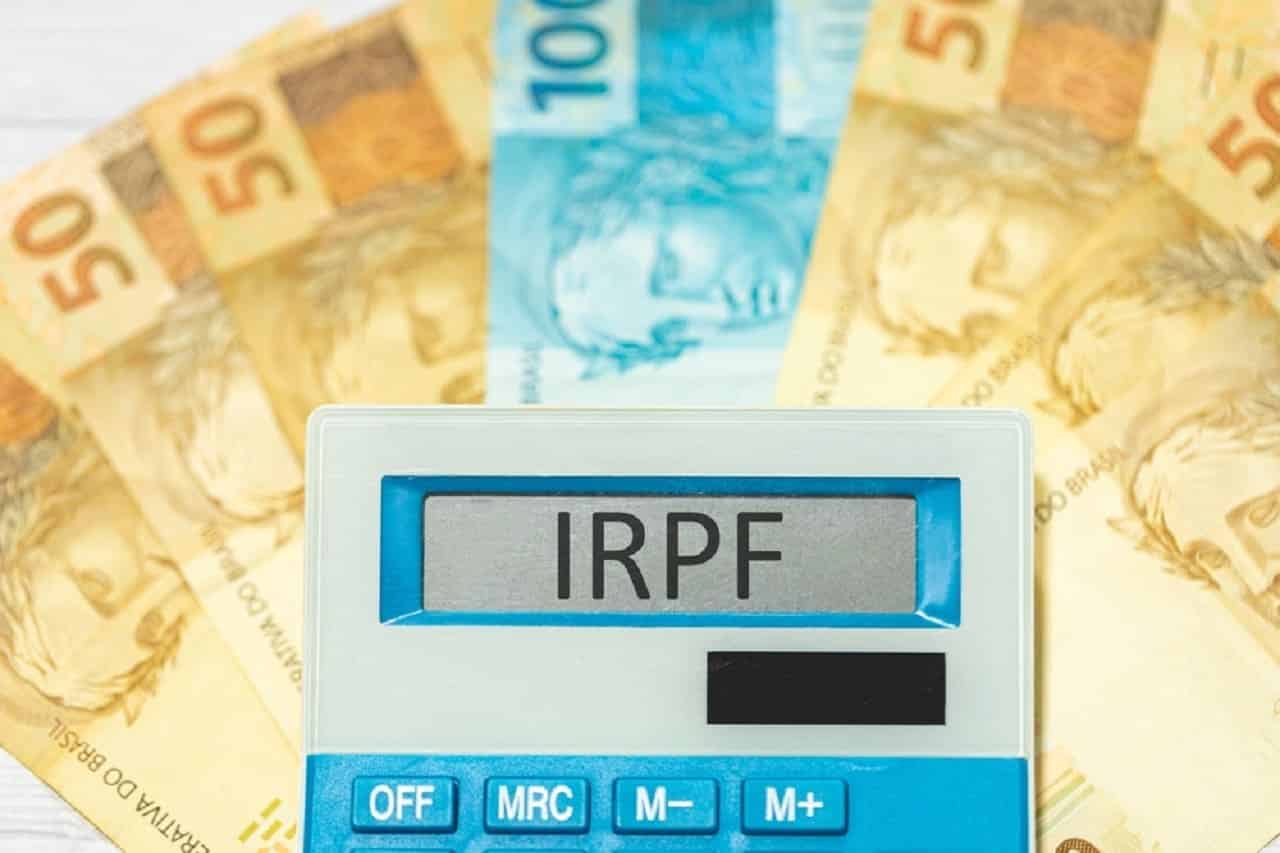 Notas de R$50 e R$100 sobre uma mesa, formando um leque. Na frente, uma calculadora com o escrito "IRPF".