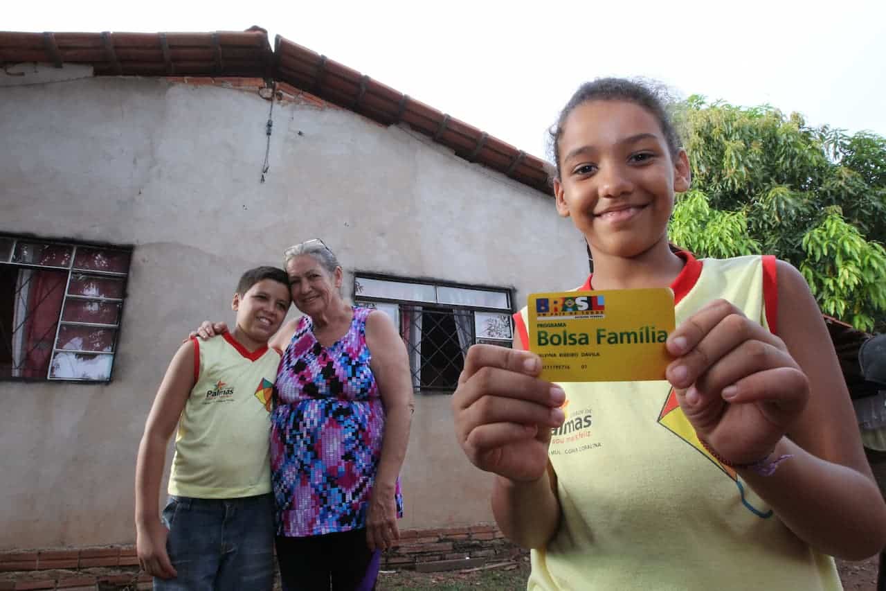 Família composta por uma senhora adulta e duas crianças em frente a uma casa com cartão Bolsa Família