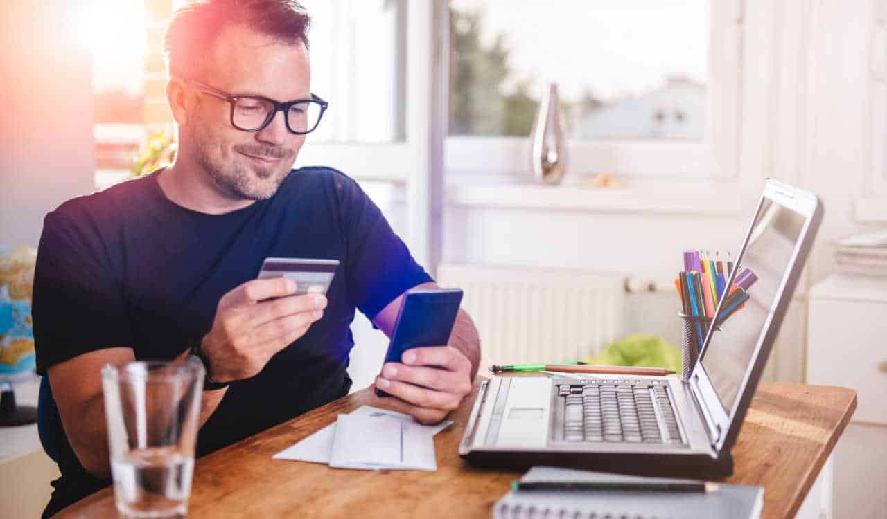 homem pedindo aumento de limite de cartão de crédito enquanto segura cartão e celular nas mãos, na sua frente está um notebookk