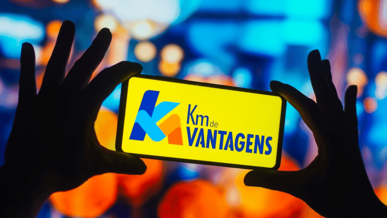 Pessoa segurando um celular que está ligado na tela em que aparece a escrita "km de vantagens" diante de um fundo amarelo