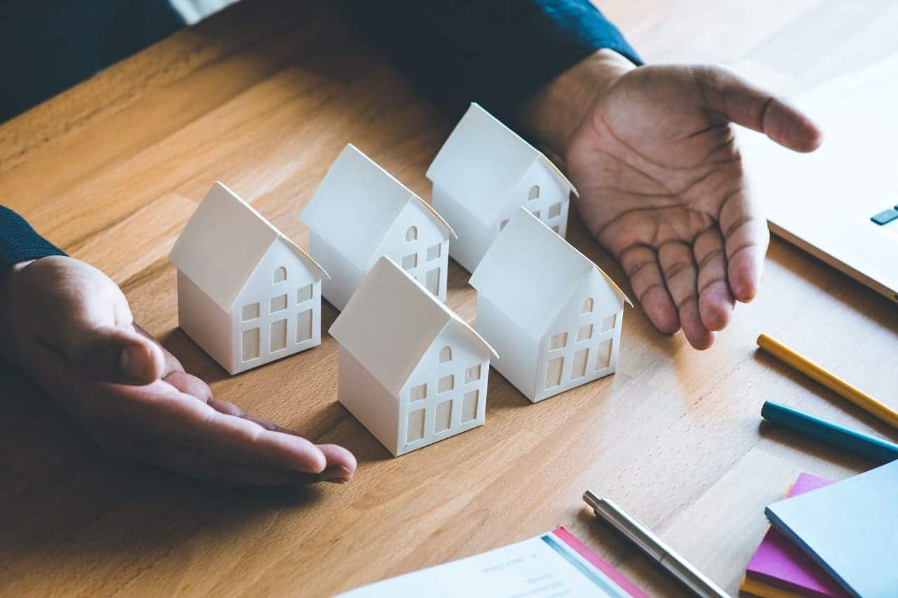 Imagem mostra uma mão masculina mostrando cinco maquetes brancas de casas sobre uma mesa