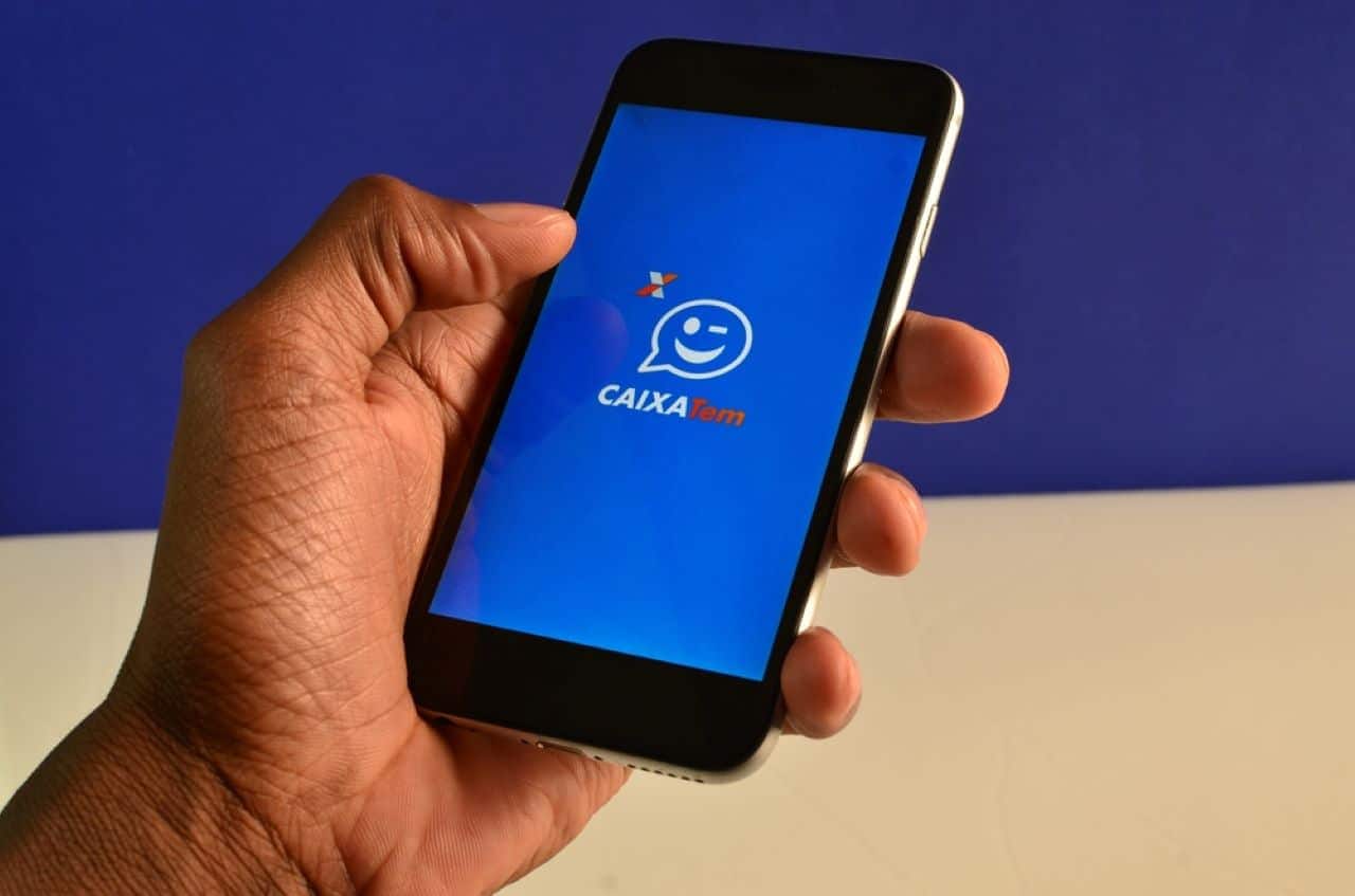 Mão masculina segurando celular com o aplicativo Caixa Tem aberto. Fundo azul e branco