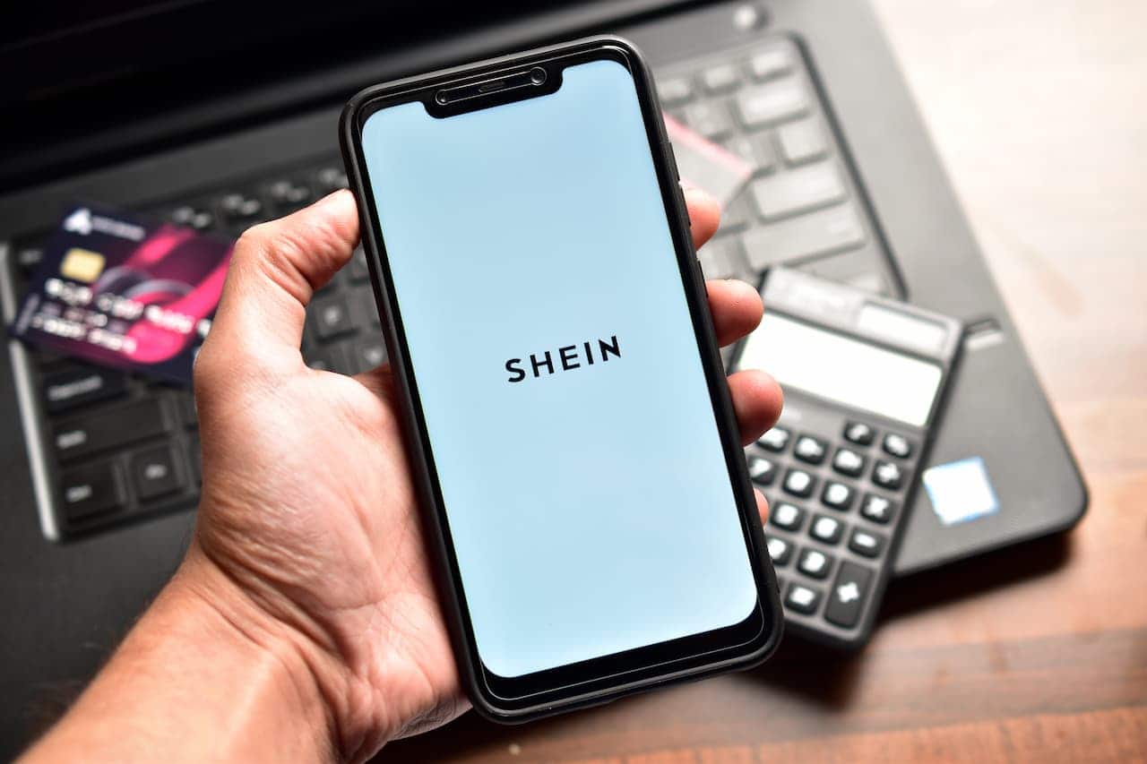 mão segurando celular com aplicativo Shein. Ao fundo, há um laptop, cartão e calculadora