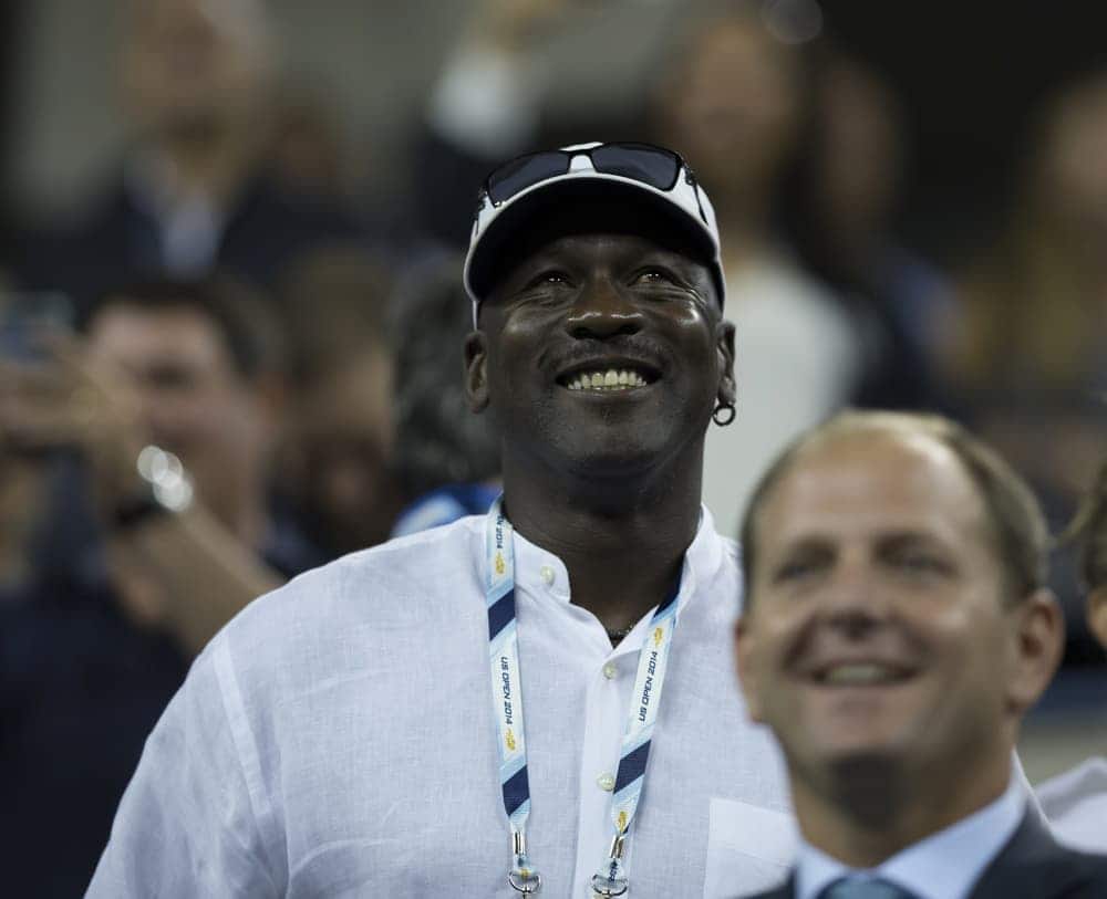 Michael Jordan de boné sorrindo. Pessoas ao fundo desfocadas.