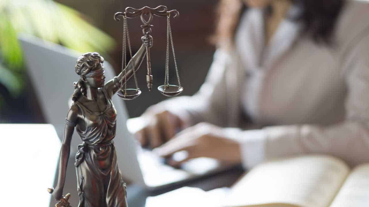 Miniatura da estátua da Justiça com mulher ao fundo enquanto digita em notebook TJ