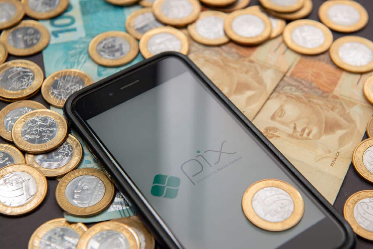 notas de cem e cinquenta reais junto com moedas de um real e um celular exibindo a logo do Pix