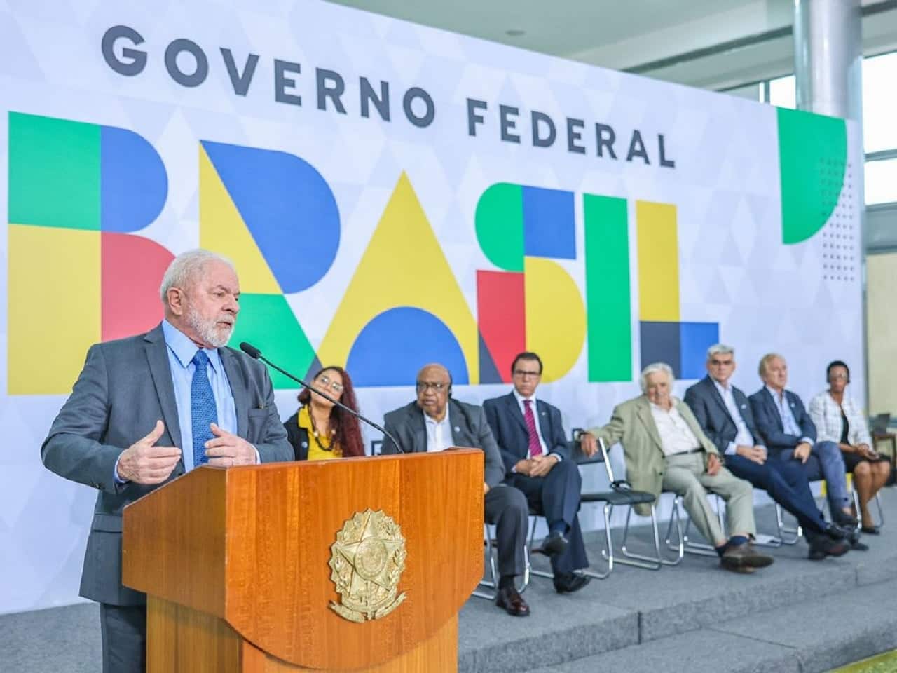 Imagem de Lula discursando. Ao fundo, um banner do Governo Federal e autoridades, todos desfocados.