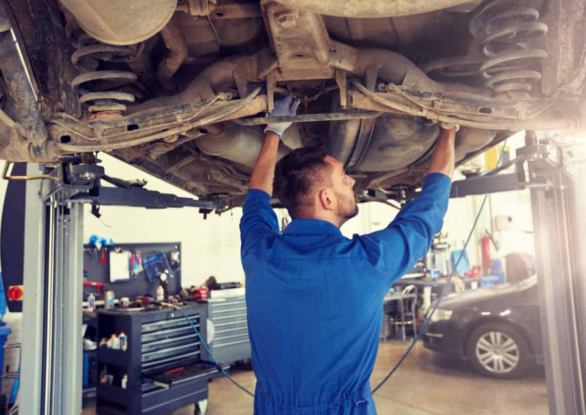 Mecânico vestido de azul em baixo de carro que está suspenso em um elevador, enquanto realiza alguma manutenção no veículo