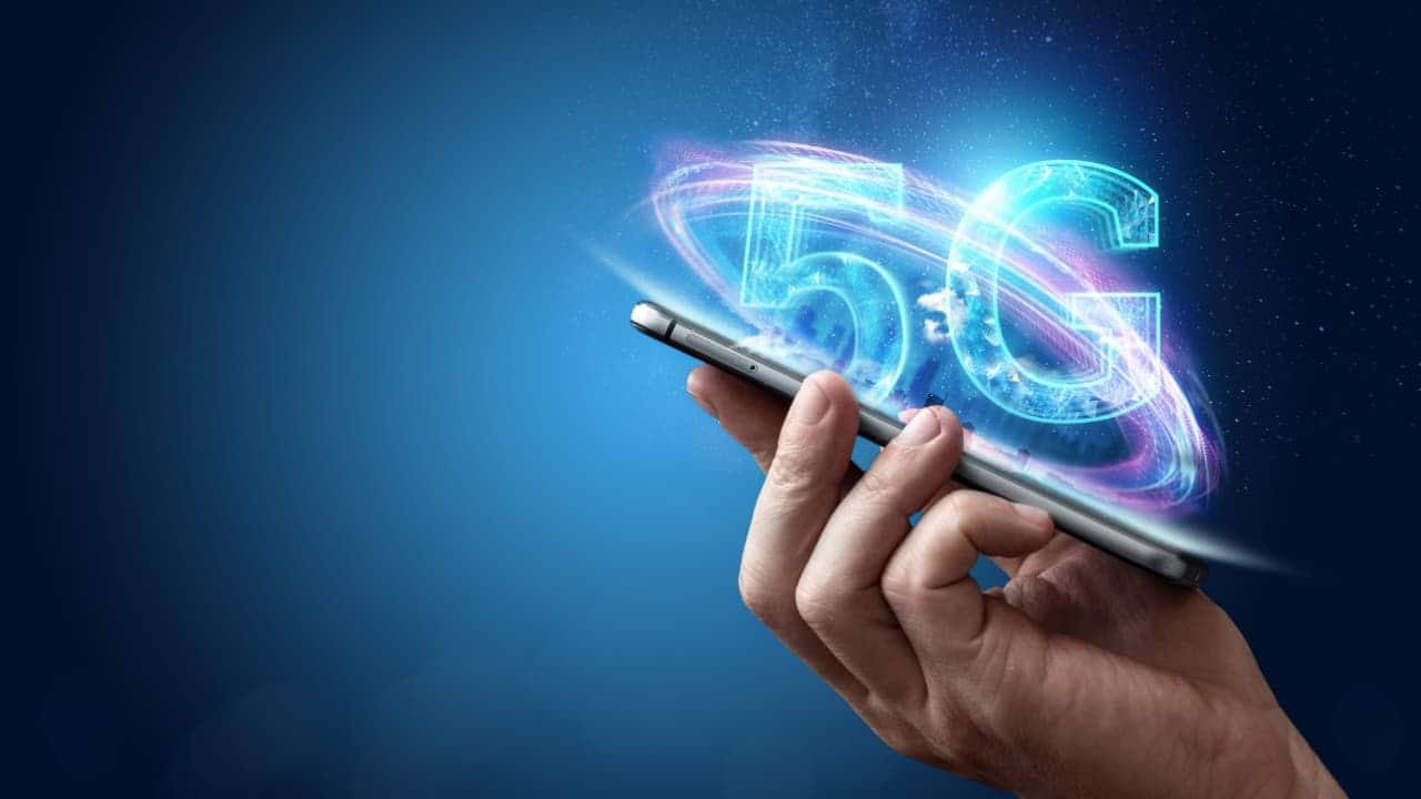 Mão segurando um celular de cuja tela sai uma animação digital do termo “5G”
