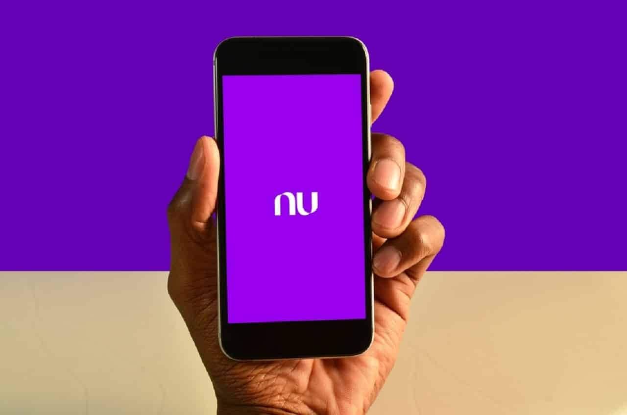 Imagem de uma mão segurando um celular com o aplicativo do Nubank aberto. O fundo é roxo e bege.