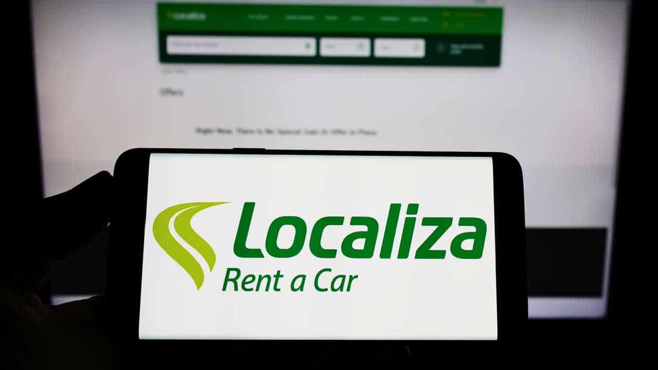 celular na mão com a logomarca da Localiza na tela inicial e, ao fundo, o site da empresa aberto na tela do computador