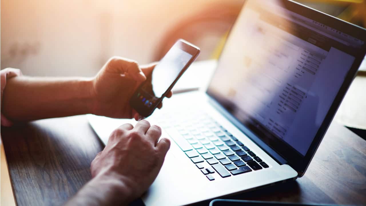 Uma pessoa cuja apenas as mãos estão amostra segurando um celular enquanto digita no computador