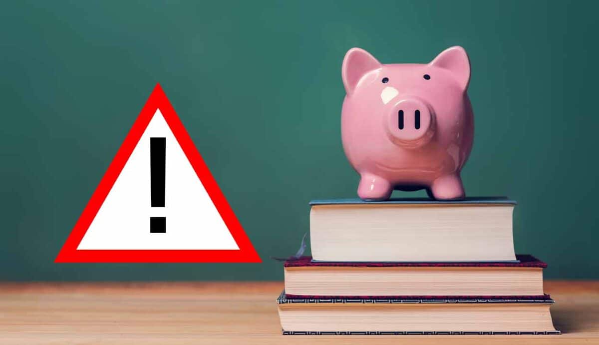 símbolo de alerta ao lado de um cofrinho de porquinho por cima de livros, ao fundo, um quadro negro.