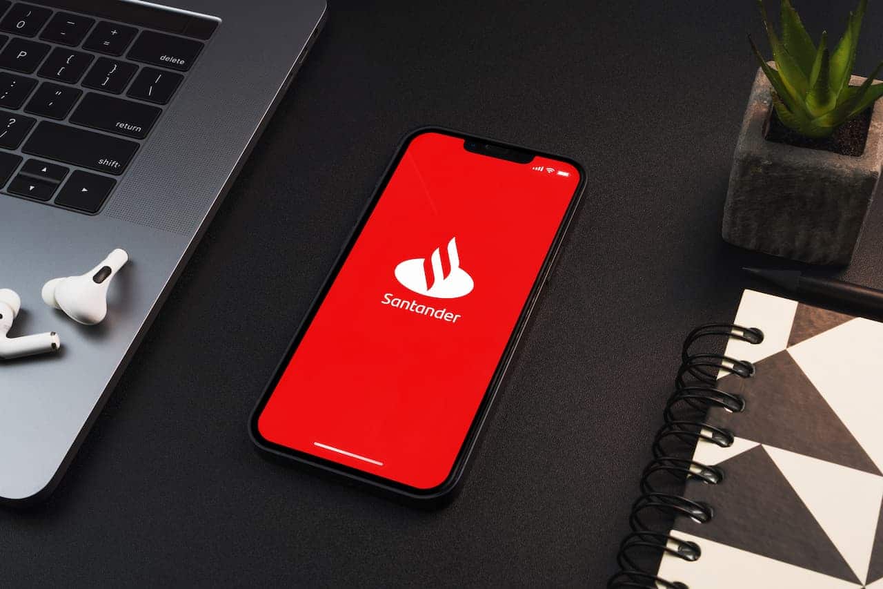 celular com a logo do Santander ao lado de laptop, fones de ouvido e caderno