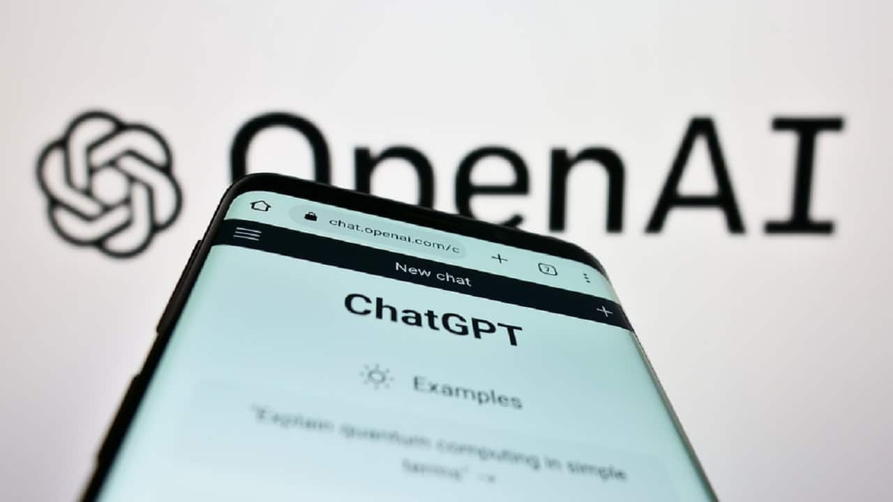 Imagem de um celular com a tela exibindo o ChatGPT. Ao fundo, a imagem da logo da OpenAI