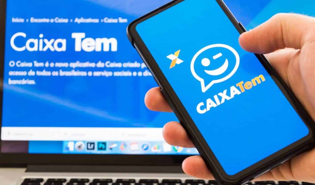 Mão segurando celular que mostra logo do aplicativo Caixa Tem. Ao fundo, tela de computador mostrando página do aplicativo no site da Caixa.