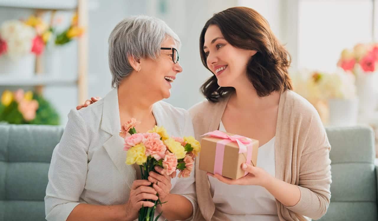 Uma mulher idosa segurando um buquê de flores e sorrindo para uma mulher mais nova segurando uma caixa de presentes