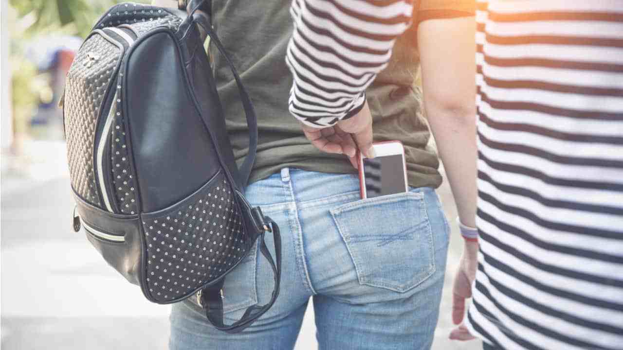 Na imagem uma mulher está na rua com o celular no bolso de trás da calça jeans e um indivíduo em furtos estica a mão para pegar o aparelho