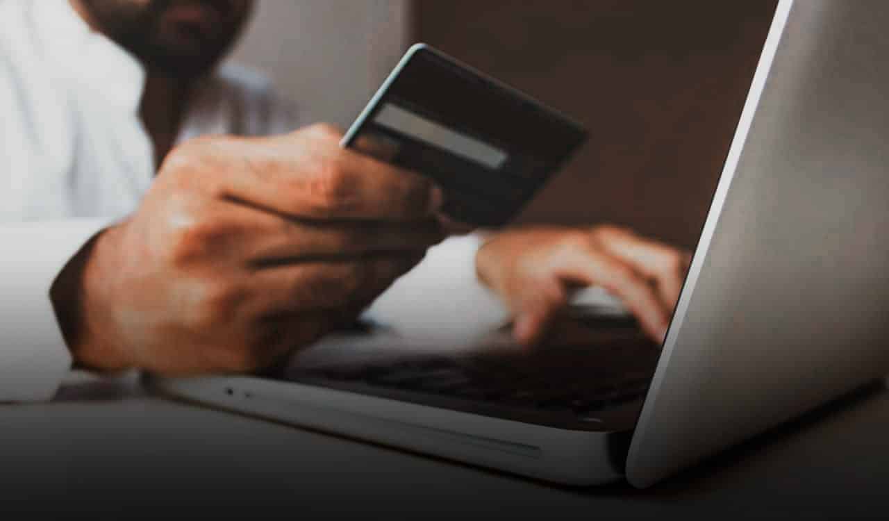 Homem digita no notebook com uma das mãos, e na outra segura um cartão de banco. fraude