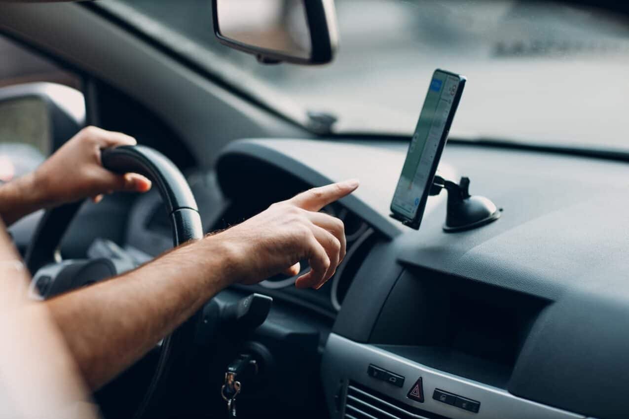 Motorista de aplicativo de transporte apontando dedo para celular enquanto segura o volante com a outra mão.